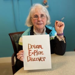 a senior woman resident of pacifica senior living mission villa holds her artwork that says dream explore discover