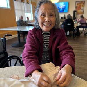 a senior asian woman, resident of pacifica senior living mission villa smiles at the camera as she folds napkins